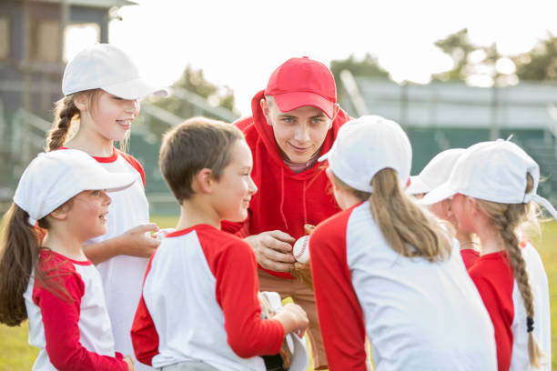 young man volunteers as coach with little league team and is teaching them about baseball during huddle - youth league imagens e fotografias de stock