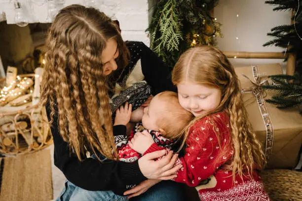 Breastfeeding baby. Mother feeding breast newborn sitting near Christmas tree in living room. Concept of lactation infant, postpartum period anywhere. Happy mom hug kids relaxing in New Year at home.