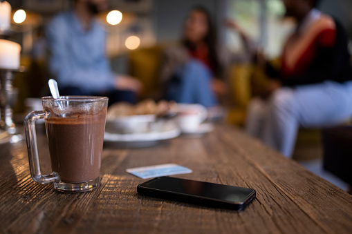 Close up view of a hot chocolate drink, smart phone and a credit card placed on a coffee table in a coffee shop with three blurred friends sitting on a sofa in the background