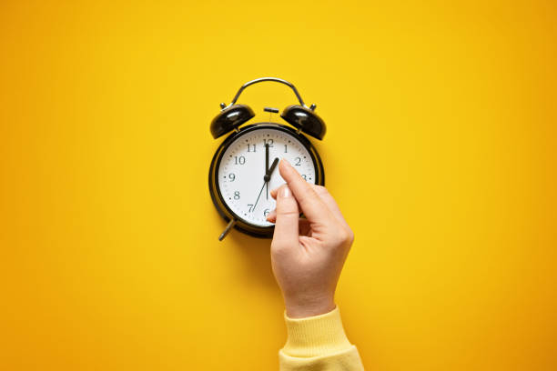 istock Daylight saving day. Fall Back. Black Alarm clock and female hands on yellow background. Daylight saving time end 1438987601
