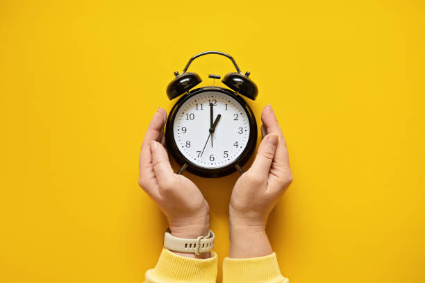istock Daylight saving day. Fall Back. Black Alarm clock and female hands on yellow background. Daylight saving time end 1438987626