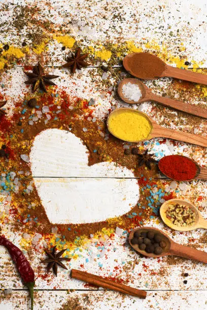 Wood spoons with turmeric, paprika, sea salt, cinnamon, anise and scattered seasoning. Set of spices on white wood background. Composition of condiment making heart shape. Cuisine and flavours concept