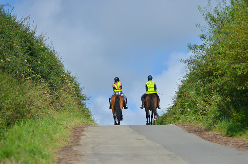 Over he hill, two women ride their horses on narrow country road both wearing brightly coloured safety gear so that they are clearly visible to other road users, keeping everyone safe.