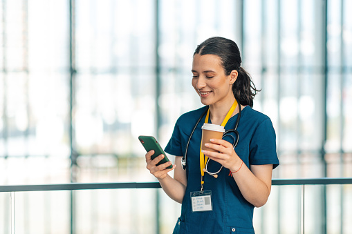 Cheerful smiling young Caucasian female nurse drinking coffee at work and surfing the net using phone