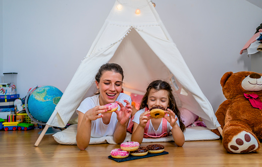 Cheerful mother and daughter eating doughnuts together in children's tent at home