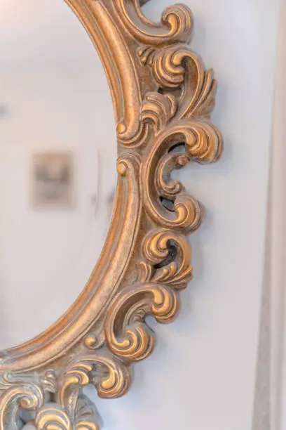 A partial view of an ellipse shaped old mirror with gold coloured and ornamented frame on a white wall.