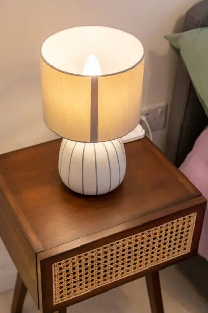 A white bedside lamp with white ceramic bottom with gray stripes and cream coloured fabric shade is on a wooden bedside table which has a drawer with a cane front. The photo is taken from an above angle, the lamp is on.