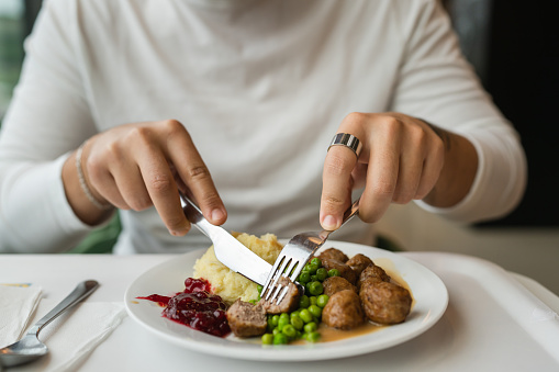 Close up on hands of unknown caucasian man sit at the table with fork and knife eat meatballs with peas and mashed potato