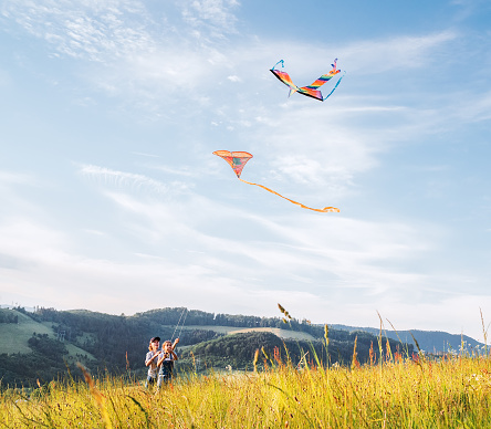 Smiling sister with launching brother with colorful kites - popular outdoor toy on the high grass hills meadow. Happy childhood moments or outdoor time spending concept image.