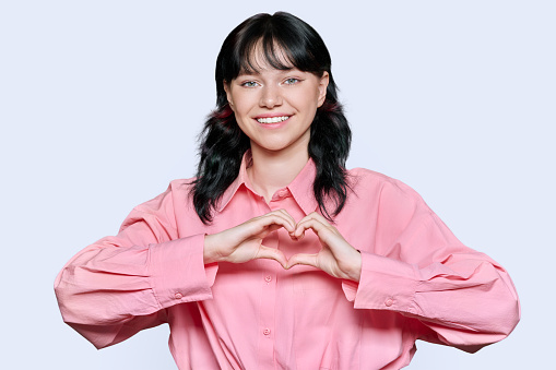 Young smiling female showing heart gesture with fingers, happy female showing love, white studio background. Signs symbols, love, emotions, romance youth concept