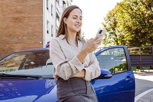 Beautiful elegant young woman leaning against her car and using a smart phone