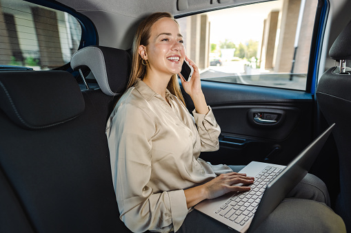 Beautiful young woman sitting on a back seat of the car and using laptop while talking on the phone