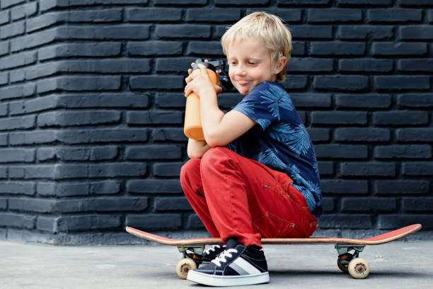 Young skater sit on skateboard, drink fresh water from reusable bottle after children training class. Active family lifestyle, outdoor recreational activities on summer holidays in city. Kids sports.