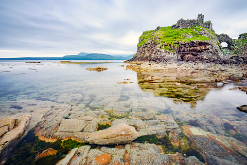 Next to the peaceful waters at low tide,,in mid summer,remains of an acient,historic Scottish castle,overlook,smooth surface of the Loch and many boulders and pebbles in seawater pools.