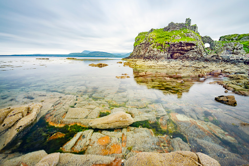 Next to the peaceful waters at low tide,,in mid summer,remains of an acient,historic Scottish castle,overlook,smooth surface of the Loch and many boulders and pebbles in seawater pools.