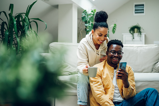 Multiracial young couple sitting on a comfortable sofa using smartphone and holding cups of hot beverages. Spouse relaxing together on cozy couch and online shopping on a mobile phone. Copy space.