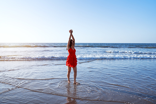 young latin woman standing on the shore of the beach in a red dress with her arms raised enjoying the sunset and the sea breeze