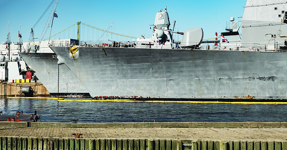 One crew member is raising two maritime flags, while the other watches or assists, on the bridge of USS Canberra (LCS 30), an Independence Class combat ship of the US Navy which is docked at Garden Island in Sydney Harbour.  She was in port for her commissioning ceremony on 22 July 2023.  As part of the ceremony, her captain was presented with a kangaroo insignia attached to all ships of the Royal Australian Navy, but decorated in the colours and design of the Stars and Stripes.  That insignia has been attached to the ship and visible on the right.  The name of the ship and serial number is visible on the orange life ring.  A large, padded chair is visible on the bridge.  This image was taken on a cloudy afternoon on 29 July 2023.