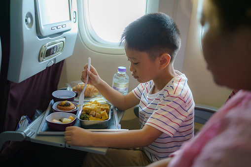Asian Child in airplane window seat. Kids flight meal. Children fly. Special inflight menu, food and drink for baby and kid. Boy eating healthy lunch in airplane. Travel and family vacation.