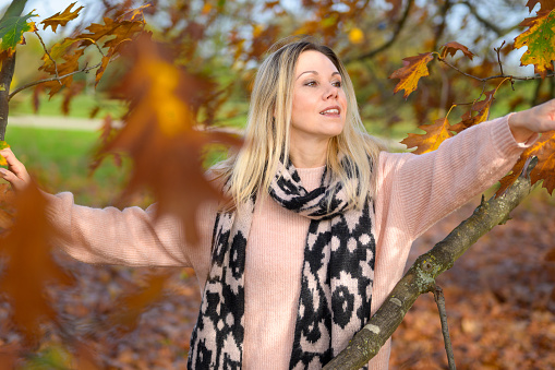 Cheerful middle-aged woman wearing a sweater and scarf is reaching for the leaf of a tree with her hand in an autumn setting
