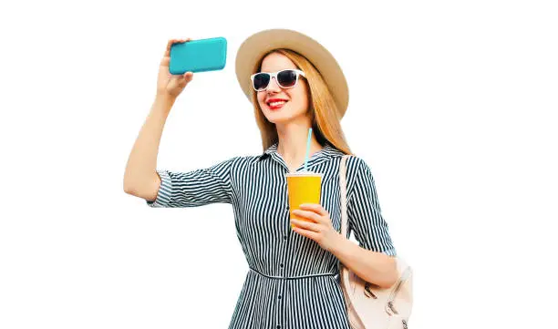 Portrait of smiling young woman taking selfie by smartphone with cup of juice wearing summer straw hat and backpack isolated on white background