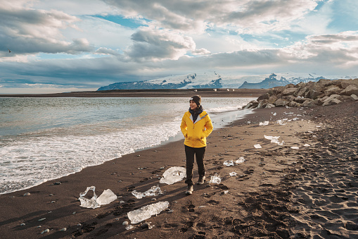 Young asian woman in yellow jacket walking on Diamond beach with iceberg melting in summer at Iceland