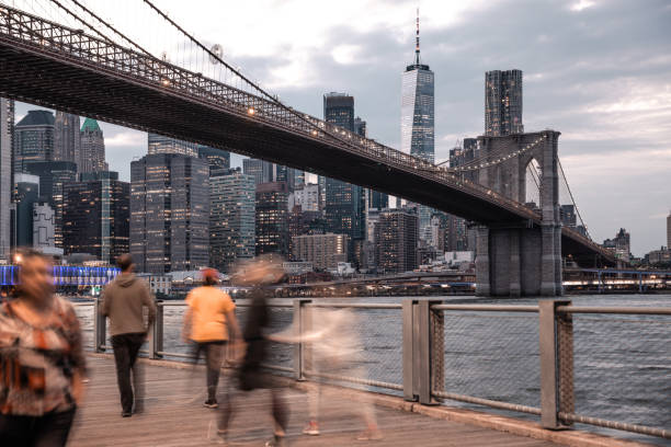 People walking on riverside walkway in Brooklyn Bridge park with Lower Manhattan downtown skyscrapers and Brooklyn Bridge on the background.