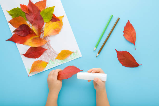 istock Little child hands gluing and creating tree shape from colorful leaves on white paper. Light blue table background. Pastel color. Making autumn decorations. Point of view shot. Closeup. Top down view. 1439147009