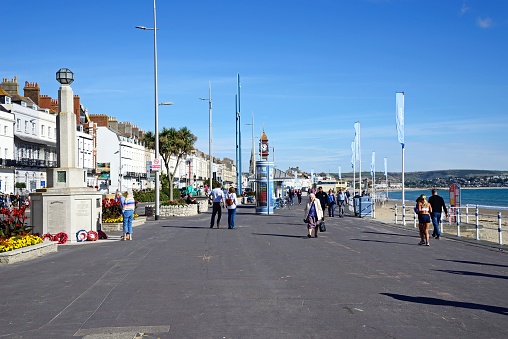 Tourists walking along the promenade with the beach to the right and promenade buildings to the left hand side, Weymouth, Dorset, UK