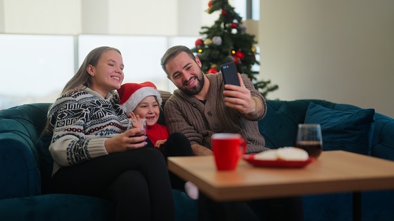 A young family is sitting in the living room at home and using smart phone to make a video call to connect with other family members or friends during Christmas.