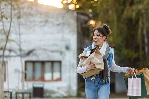 Copy space shot of cheerful young brunette woman walking down the city street, carrying takeaway food order and shopping bags. She is looking away, smiling joyfully and contemplating.