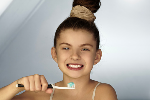 A pretty 9 year old girl while brushing her teeth, holding a toothbrush in her hand and smiling. She is looking straight ahead and holding a toothbrush with toothpaste.