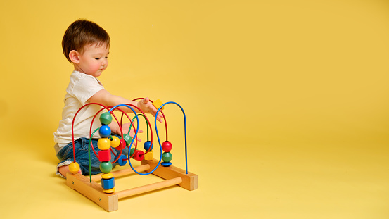 Toddler baby is playing logic educational games on a studio yellow background. Happy child play with educational toy, learning logic, copy space. Kid aged one year and four months