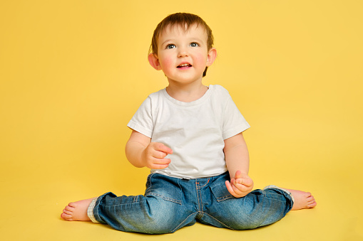 Portrait of a happy toddler baby in full length on a studio yellow background. Smiling child sitting on the floor in a white t-shirt and blue jeans, copy space. Kid aged one year and four months