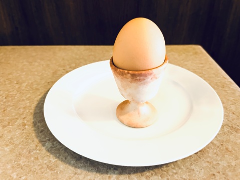 Horizontal closeup photo of an organic hard boiled egg in a ceramic eggcup on a round white plate on a kitchen table.