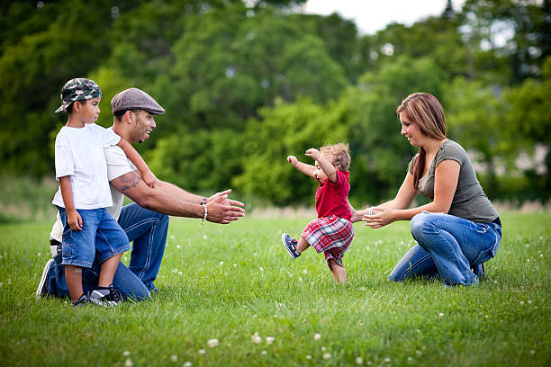 Toddler Learning to Walk with Happy Family Outside A happy, young multiracial family of four spending time together outside while their one year old son learns to walk. first steps stock pictures, royalty-free photos & images