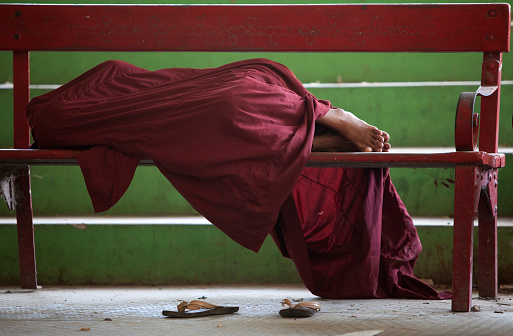 A monk uses his robes to cover himself completely while he sleeps on a wooden bench.