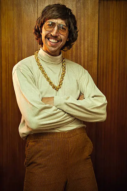 A smiling goofy man from the 1970s - 1980s poses for a picture in his turtleneck, high waisted elastic corduroy pants, and his fancy gold chain necklace .  Classy tinted glasses and suave mustache.  Wood panel wall in the background.