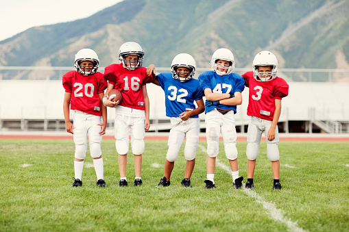 A low-angle, wide shot of a male football coach and a group of children wearing sports clothing, football boots and sports bibs on a football pitch in the North East of England. The coach stands and motivates his students as they run and jump over agility hurdles.