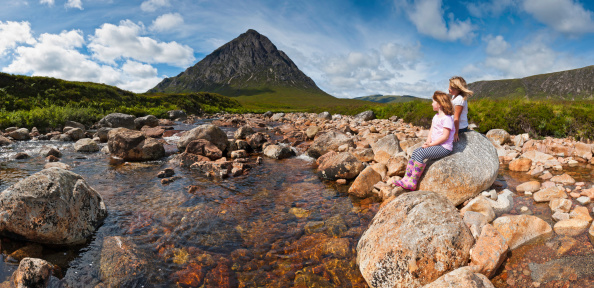 Two young children sat on a rock beside a clear mountain stream looking at the iconic rocky pyramid of Buachaille Etive Mor, Glen Coe, deep in the picturesque Highlands of Scotland. ProPhoto RGB profile for maximum color fidelity and gamut.