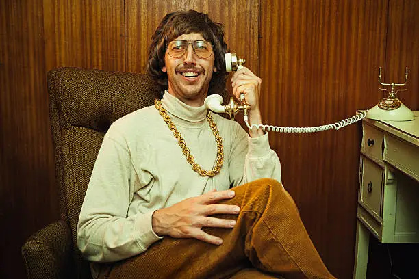 A smiling goofy man from the 1970s - 1980s relaxes as he talks on his vintage rotary telephone. He's wearing a turtleneck, high waisted elastic corduroy pants, and his fancy gold chain necklace .  Classy tinted glasses and suave mustache.  Wood panel wall in the background.