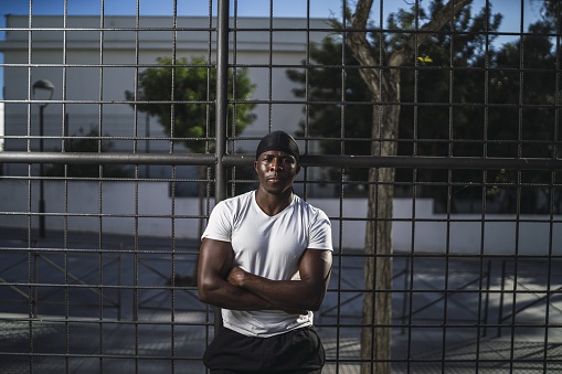 A shallow focus shot of an African-American male in a white shirt leaning on a fence with arms crossed