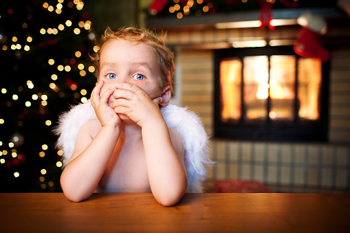 Little angel holding his hand against the mouth sitting in front of Christmas tree and fireplace.