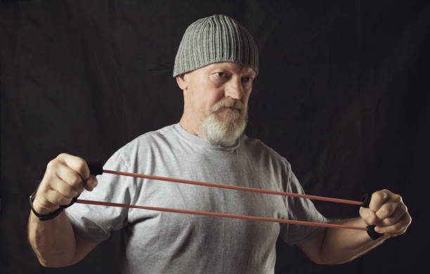 Fitness, an elderly stern man stretches a rubber expander Fitness, an elderly stern man stretches a rubber expander. senior bodybuilders stock pictures, royalty-free photos & images