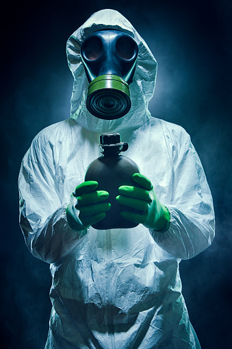 A man in a Hazmat chemical protection suit and gas mask stands in the midst of gas, smoke, or fog, holding a small canteen of rationed water.  Vertical.