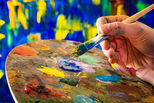 Closeup of a painter hand holding a paintbrush picking blue and yellow oil paint from the palette to paint a canvas. Predominant colors are blue and yellow.