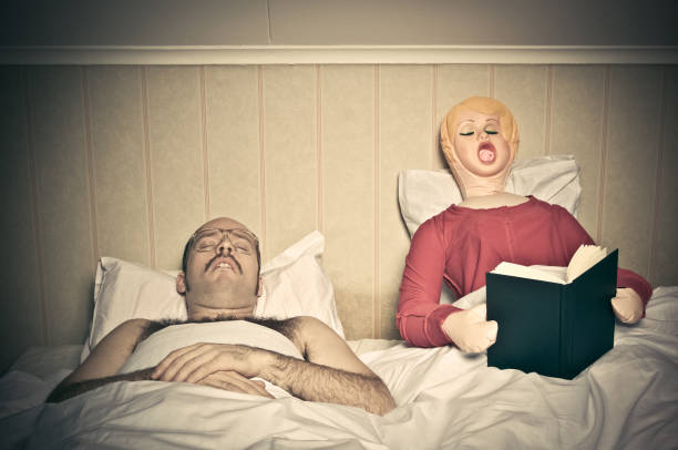 Bed Time Routines Nerdy looking guy sleeps while blow up doll reads a book in bed. blow up doll stock pictures, royalty-free photos & images