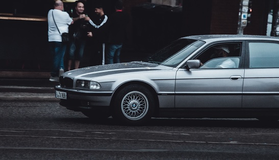 Berlin, Germany – August 20, 2022: A closeup shot of a gray BMW vehicle on the street road in Berlin, Germany, with people toasting with glasses in the background