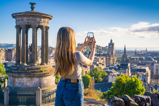 Student girl shooting photograph with smartphone in Edinburgh at sunset from Calton Hill with skyline of Edinburgh, capital city of Scotland UK United Kingdom
