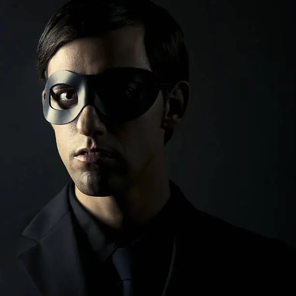 young man wearing all black - shirt, suit and tie, also wearing a dark mask an staring at camera with a sinister look